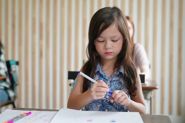 Disegno del bambino della ragazza in età prescolare con la matita di colore su carta bianca sul tavolo in aula con gli amici