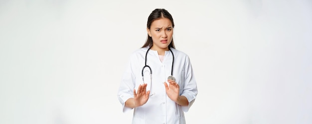 Disgustato dottoressa asiatica che rifiuta di dire di non guardare con antipatia rifiutando di stare in piedi sgradevole in uniforme medica su sfondo bianco