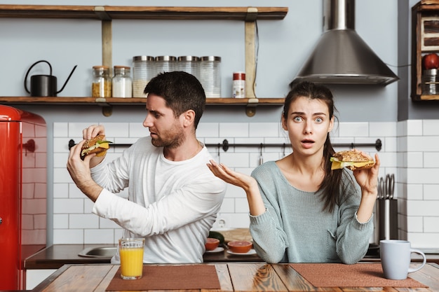 Disgustato giovane coppia seduta in cucina durante la colazione a casa, con in mano panini