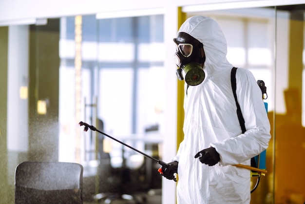 Disinfezione dell'ufficio per prevenire COVID-19, uomo in tuta protettiva ignifuga con prodotti chimici spray.