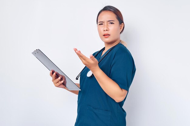 Dispiaciuta e infastidita giovane infermiera donna asiatica che indossa uniforme blu con uno stetoscopio che legge sciocchezze su un blocco appunti isolato su sfondo bianco Concetto di medicina sanitaria