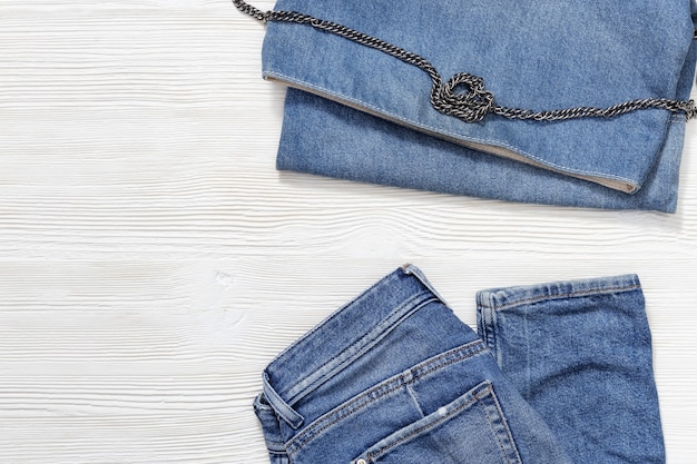 Disposizione piana degli abiti di moda casual delle donne su fondo di legno bianco, blue jeans e borsa in denim. Abbigliamento femminile con spazio di copia.