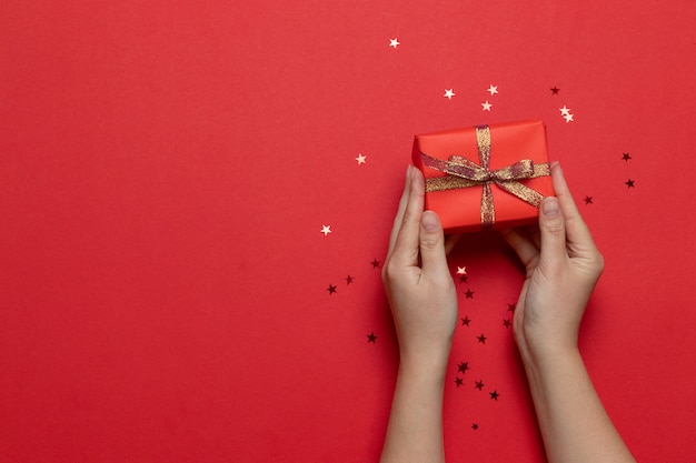 Disposizione piana delle mani della donna che tengono il contenitore di regalo di sorpresa avvolto e decorato con l'arco con le stelle d'oro su fondo rosso. Compleanno, San Valentino, Natale, Capodanno. Stile piatto laico