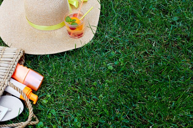 Disposizione piana di vacanze estive luminose all'aperto o concetto di stile di vita di viaggio con borsa di paglia, cappello, infradito, limonata e spray solare su sfondo di erba, vista dall'alto, spazio per la copia.