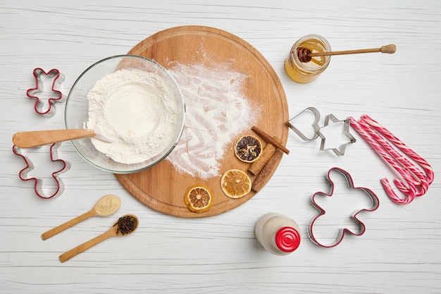 Disposizione piatta degli ingredienti per la preparazione di pasta e biscotti di panpepato, stampi da taglio, bottiglia con latte vegetale, lecca lecca di Natale, barattolo di vetro con miele e ciotola con farina su una tavola di legno rotonda