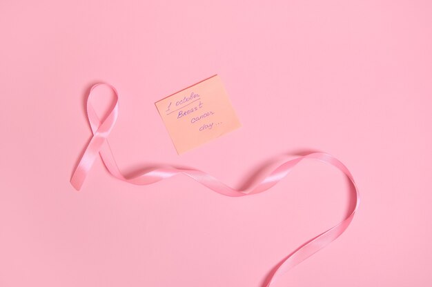 Disposizione piatta di un lungo nastro rosa con un'estremità infinita e una nota di carta con scritta 1 ottobre Giornata di sensibilizzazione sul cancro al seno, isolata su sfondo rosa con spazio per il testo