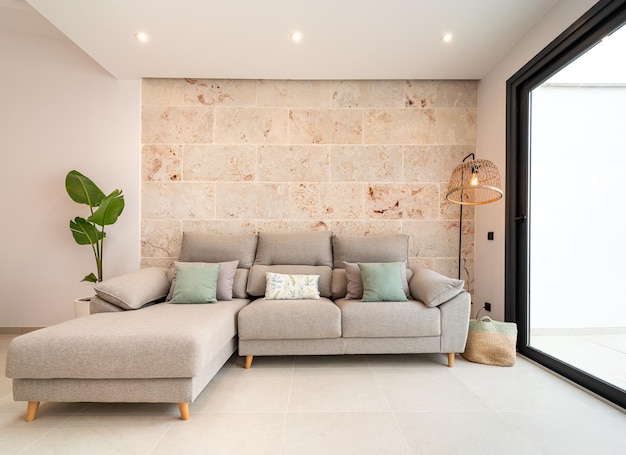 Divano del soggiorno grigio chiaro decorato con cuscini, una lampada e una pianta davanti a un muro di pietra
