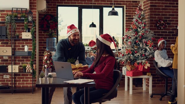Diverse persone che fanno regali e regali ai colleghi, celebrando le vacanze invernali con decorazioni festive nell'ufficio dell'azienda. I colleghi con i cappelli di Babbo Natale si sentono allegri durante il periodo natalizio.