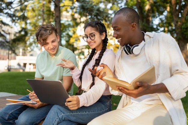 Diversi studenti felici che studiano insieme nel parco si preparano per il test e guardano lo schermo del laptop