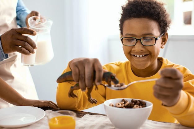 Divertendosi. Piacevole ragazzo allegro che gioca con un dinosauro giocattolo e lo nutre di cereali mentre suo padre gli versa un bicchiere di latte