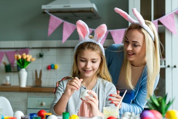 Divertente, allegra, gioiosa mamma che insegna, allena la sua piccola, carina, piccola, piccola figlia disegna, dipinge, decora le uova di Pasqua, insieme indossa le orecchie di coniglio, si prepara per la Pasqua