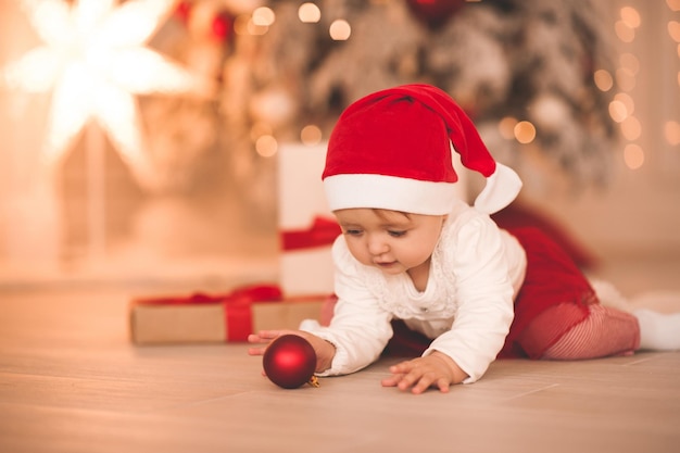 Divertente bambina di 1-2 anni che indossa un abito di Babbo Natale che gioca con le decorazioni di Natale sdraiato sul pavimento in camera sotto il primo piano dell'albero di Natale. Stagione delle vacanze invernali. Infanzia.