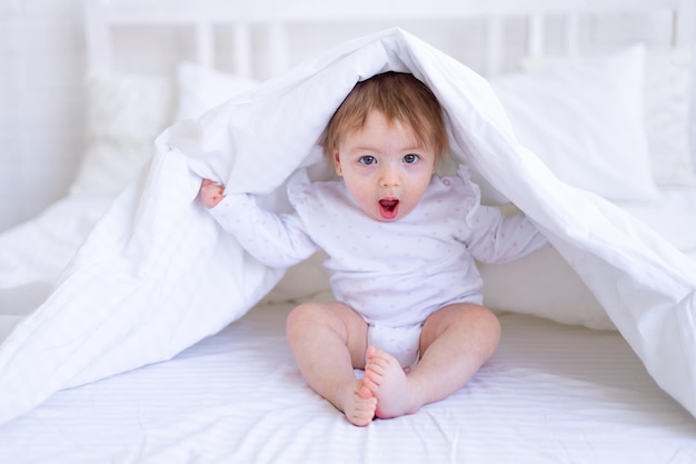 Divertente bambina sorpresa guarda fuori da sotto la coperta sul letto in abiti bianchi con la bocca aperta un bambino piccolo su un letto di cotone a casa si è svegliato la mattina dopo aver dormito
