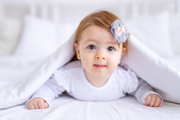 Divertente bambina sorpresa guarda fuori da sotto la coperta sul letto in abiti bianchi con la bocca aperta un bambino piccolo su un letto di cotone a casa si è svegliato la mattina dopo aver dormito