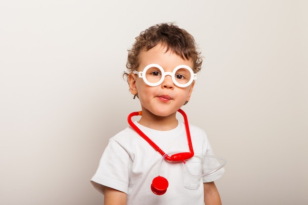 Divertente bambino riccio in bicchieri con uno stetoscopio sul collo gioca un medico. Ritratto più ampio di un bambino che gioca la professione di medico.