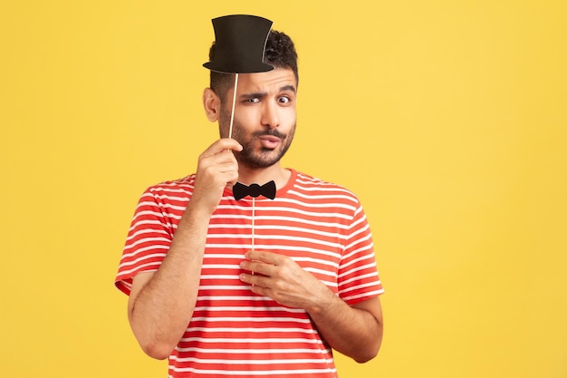 Divertente bell'uomo con la barba in camicia a righe che prova le maschere di carta cappello e farfallino sul bastone, preparandosi per una festa di fantasia, mascherata. Studio indoor girato isolato su sfondo giallo