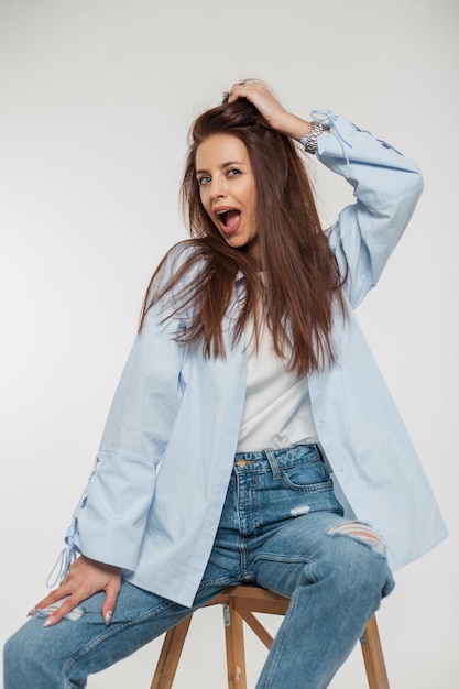 Divertente bella donna alla moda con emozioni di sorpresa e gioia in abiti in denim alla moda con camicia e jeans si siede e si diverte su uno sfondo bianco in studio