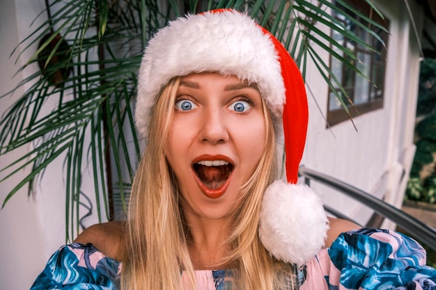 Divertente donna bionda scioccata che indossa un cappello da Babbo Natale che guarda nella fotocamera con occhi sorpresi e un mou aperto