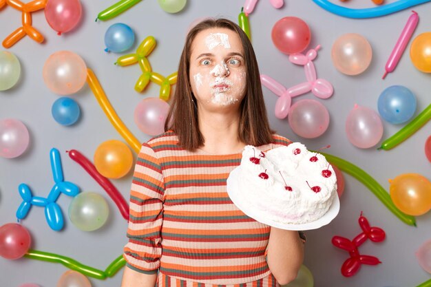 Divertente donna pazza in abito a righe in piedi contro il muro grigio decorato con palloncini colorati ragazza di compleanno con crema torta sul viso che attraversa gli occhi