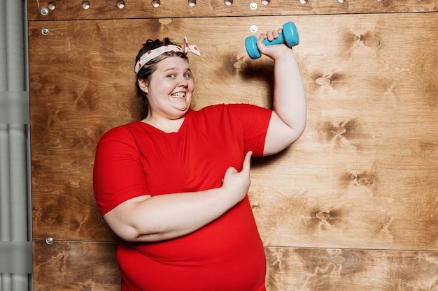 Divertente donna sovrappeso vestita con abiti sportivi e con una benda sulla testa si allena con manubri sullo sfondo di una parete di legno