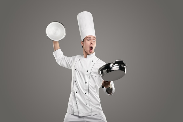 Divertente eccitato giovane chef maschio in uniforme bianca e cappello che tiene il coperchio in mano alzata e guarda con stupore la padella, mentre cucina il cibo nella cucina del ristorante isolato su sfondo grigio