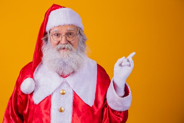 Divertente felice eccitato vecchio barbuto Babbo Natale faccia indossando il costume guardando la telecamera che mostra le dita puntate da parte pubblicità promozione natalizia, annuncio di sconto Natale Capodanno isolato su giallo