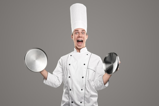 Divertente giovane chef maschio in uniforme bianca e cappello che guarda l'obbiettivo con una smorfia stupita mentre tiene in mano coperchi di padella di metallo su sfondo grigio