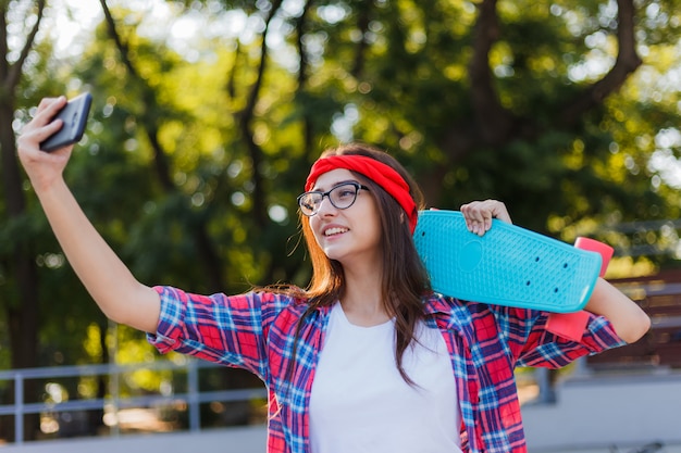 Divertente giovane donna in occhiali e camicia a quadri rossa. Skatepark della donna n dei pantaloni a vita bassa mentre fa selfie con lo skateboard nello skatepark al giorno luminoso soleggiato. Divertimento estivo