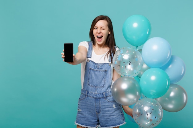 Divertente giovane donna lampeggiante tenere il telefono cellulare con schermo vuoto vuoto che celebra con palloncini colorati isolati su sfondo blu turchese parete. Festa di compleanno, concetto di emozioni della gente.