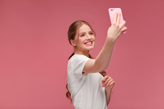 Divertente giovane donna rossa che fa selfie ragazza sorridente che indossa una maglietta bianca che tiene smartphone rosa ma...