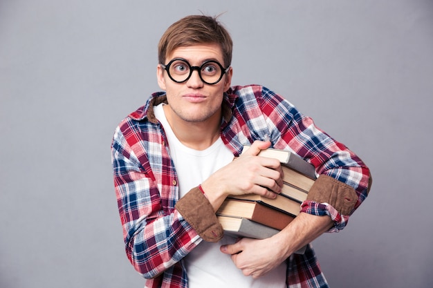 Divertente giovane maschio comico con occhiali rotondi e camicia a scacchi che tiene libri su un muro grigio