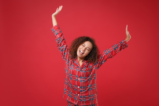 Divertente giovane ragazza afroamericana in pigiama homewear che riposa a casa isolata su sfondo rosso. Rilassatevi il concetto di stile di vita di buon umore. Mock up spazio di copia. Tenendo gli occhi chiusi, alzando le mani allargando.