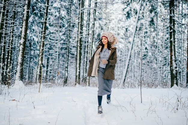 Divertente giovane ragazza attraente con un turbante dalla sciarpa che corre tra gli alberi nella foresta di neve