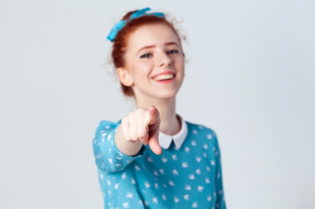 Divertente ragazza allo zenzero in abito azzurro con dito puntato verso la telecamera e sorriso a trentadue denti