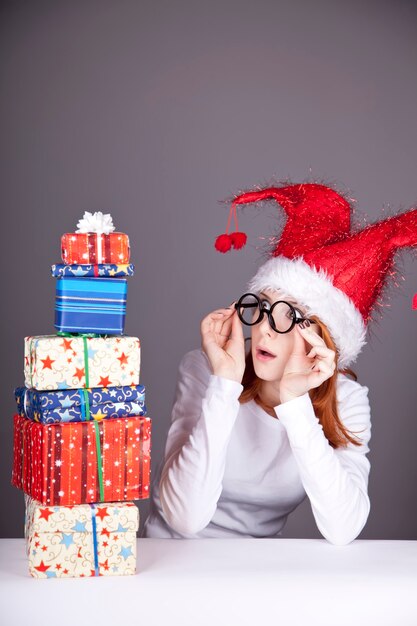 Divertente ragazza dai capelli rossi in cappello di Natale e bicchieri con scatole regalo.