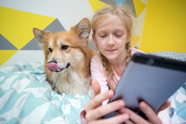 Divertente ragazza e cane corgi sul letto nella stanza dei bambini giocano sul tablet