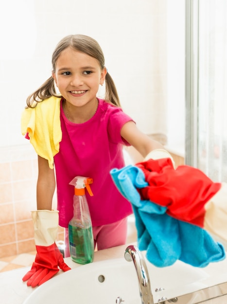 Divertente ragazza sorridente che lucida lo specchio in bagno mentre pulisce la casa