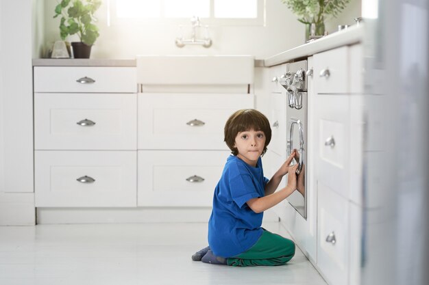 Divertente ragazzino ispanico che sembra sorpreso dalla telecamera mentre guarda la torta che cuoce nel forno, accovacciato in cucina. Bambini, concetto di cucina. Vista laterale