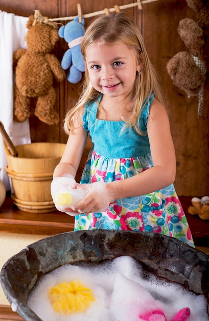 Divertente sessione fotografica domestica Una bambina curiosa lava gli orsi in un'antica bacinella di rame con acqua e schiuma di sapone a casa