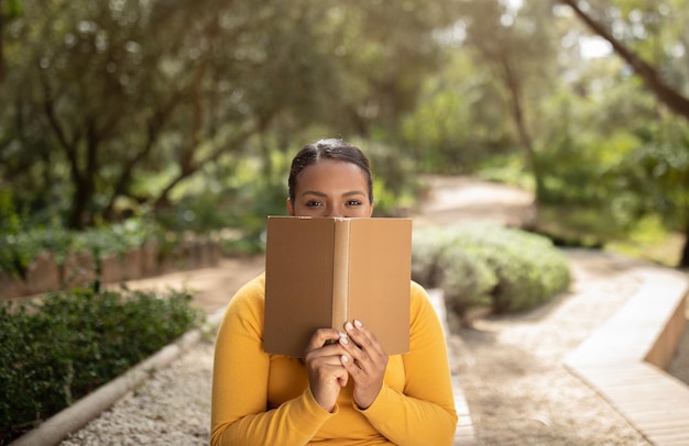 Divertente studentessa brasiliana che dà una occhiata al libro seduto nel parco che si prepara per le lezioni all'aperto