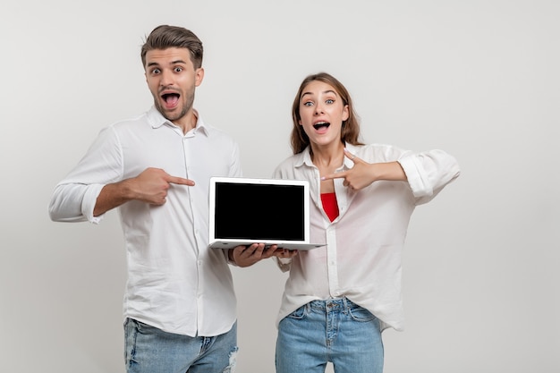 Divertenti amici di coppia che puntano il dito indice sul computer portatile con schermo vuoto isolato su bianco