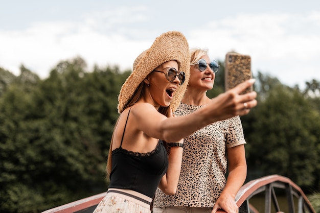 Divertenti due donne mamma e figlia con emozioni felici sul viso in occhiali da sole con un cappello di paglia in abito estivo alla moda fanno selfie al telefono e viaggiano durante le vacanze estive