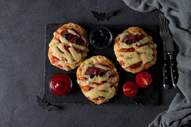 Divertenti mini pizze mummie con salsiccia e formaggio per la festa di Halloween servite con ketchup di olive nere e pomodori su sfondo scuro