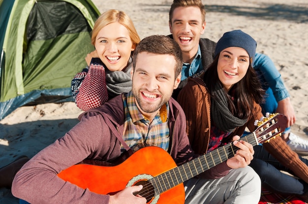 Divertirsi insieme. Vista dall'alto di quattro giovani felici seduti vicino alla tenda insieme mentre un giovane bell'uomo suona la chitarra e sorride