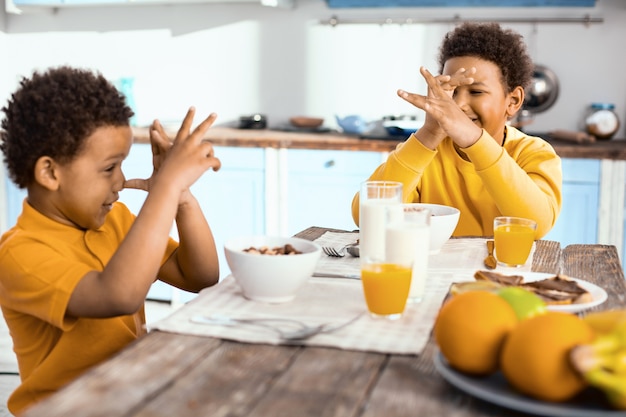 Divertirsi. Ragazzini allegri seduti al tavolo, che si guardano l'un l'altro con il naso e ridono mentre fanno colazione