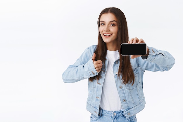 Divertito ed entusiasta studentessa di bell'aspetto in giacca di jeans che mostra lo schermo dello smartphone che tiene il telefono cellulare in orizzontale e fa il pollice in su consiglia una nuova fantastica applicazione o gioco