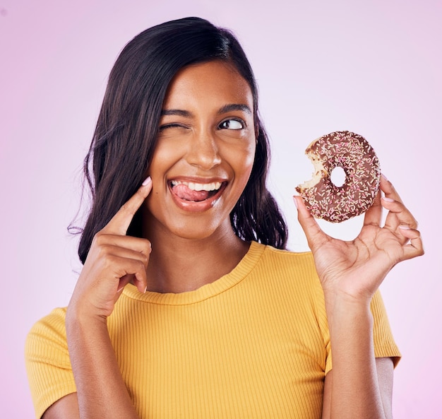 Dolce ciambella e occhiolino con donna in studio per spuntino dietetico e felicità Cibo zuccherato e sorriso con femmina che mangia dolcetto al cioccolato isolato su sfondo rosa per nutrizione giocosa e brama