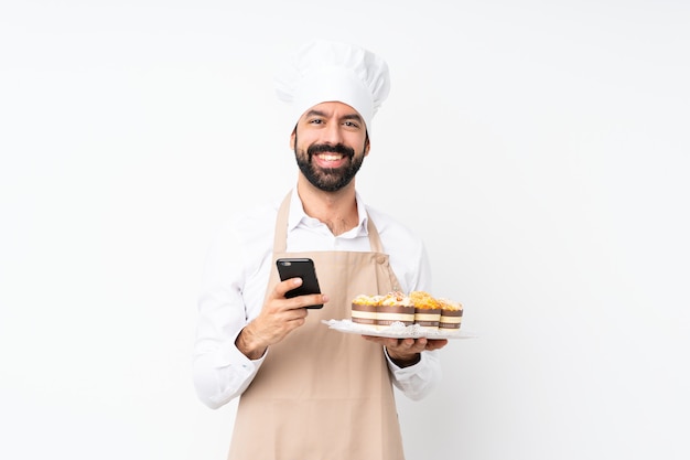 Dolce del muffin della tenuta del giovane sopra la parete bianca isolata che invia un messaggio con il cellulare