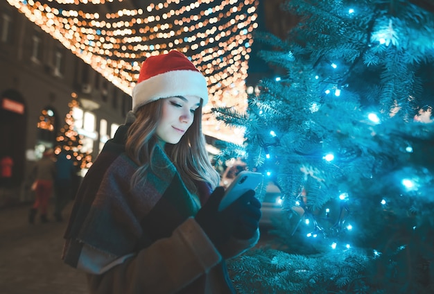 Dolce donna in un cappello di Natale si trova su un albero in una strada decorata in vacanza e utilizza uno smartphone