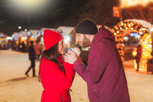 Dolce ritratto di una coppia di innamorati che si scalda le mani, tenendo in mano una tazza di bevanda calda. Momenti felici della coppia sposata all'aperto al mercatino di Natale